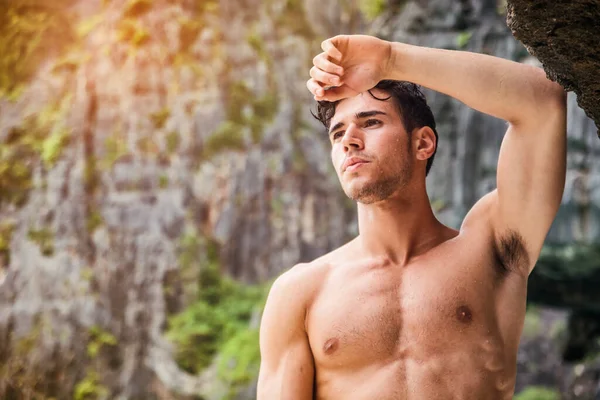 Half body shot of a handsome young man standing on a beach in Phuket Island, Thailand, shirtless wearing boxer shorts, showing muscular fit body, looking away to a side