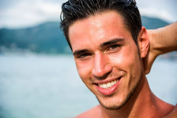 Attractive young man in the sea getting out of water with wet hair, looking in camera wiht a smile