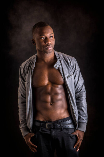 Handsome shirtless muscular black young mans with jacket open on naked torso, looking at camera, on dark background in studio shot