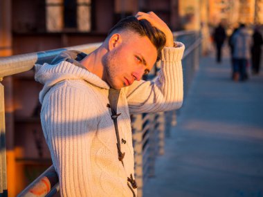 Yakışıklı genç adam, günbatımında bir kaldırımda duruyor şık bir kazak ya da hırka giyiyor.