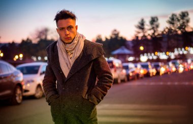 Yakışıklı, modaya uygun genç bir adam, gece vakti şehrin kaldırımında duruyor. Şık bir kışlık palto ve eşarp giyiyor.