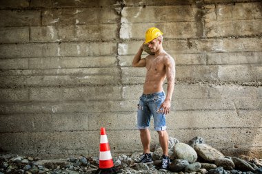 Seksi, kaslı inşaat işçisi, üstsüz, açık havada çalışıyor, kot pantolon giyiyor ve sarı şapka takıyor.