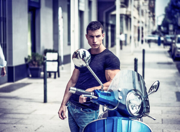 Young athletic man standing by his classic Italian scooter on empty street in sunny city.