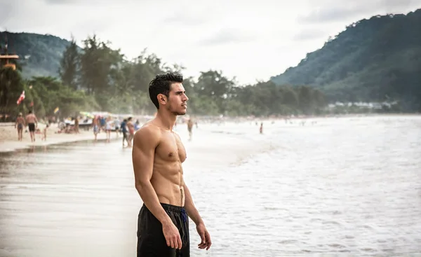 Half body shot of a handsome young man standing on a beach in Phuket Island, Thailand, shirtless wearing boxer shorts, showing muscular fit body