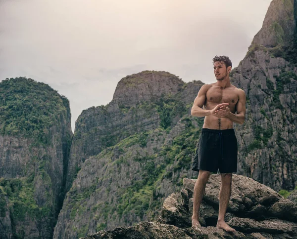 Half body shot of a handsome young man standing on a rock in Phuket Island, Thailand, shirtless wearing boxer shorts, showing muscular fit body