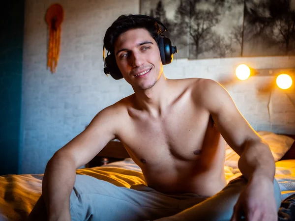 彼のベッドの上に一人で横たわっているヘッドフォンを使用して魅力的な若い男 音楽を聴き 楽しい時間を持っています カメラを見て — ストック写真