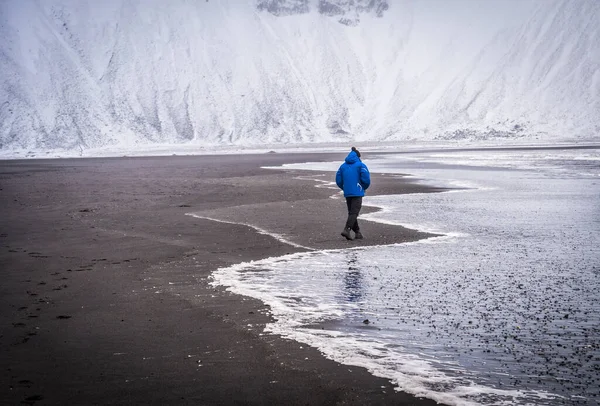 アイスランドの雪の山の近くに波を振って海に沿って歩く青い暖かいジャケットの認識できない男性探検家の完全な体の背景色 — ストック写真