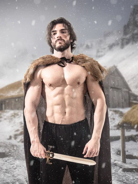 Bearded masculine Northern warrior with muscular shirtless core wearing fur coat and holding sword against traditional house in snowfall