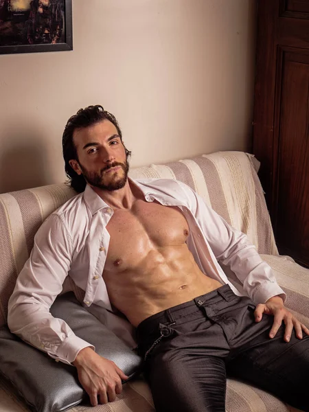 Handsome muscular young man at home laying on couch with shirt open on naked torso, an expression of tranquility, looking at camera, in a health and fitness concept