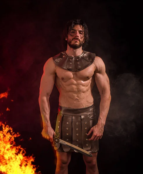 Young handsome muscular man posing shirtless in roman or spartan gladiator costume in studio. A handsome and well-built man strikes a pose while shirtless, dressed in a Roman or Spartan gladiator costume, set against a black backdrop.