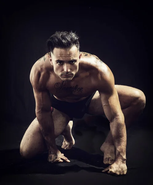 A man with a tattoo on his chest in a Tarzan pose, kneeling and crouching while looking at camera, almost ready to spring