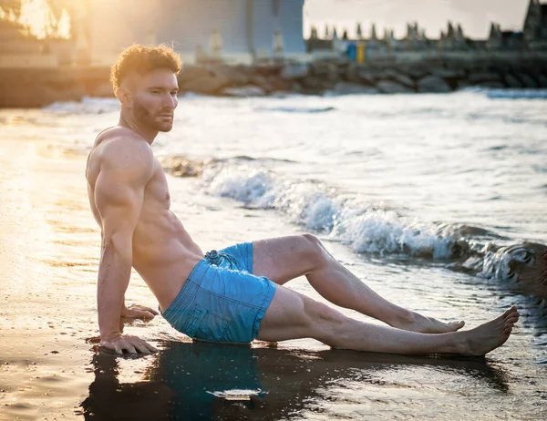 夜明けに海の隣のビーチに座っているシャツのない筋肉の男 — ストック写真
