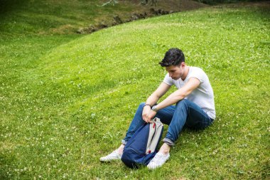 Sırt çantasıyla çimenlerde oturan genç bir adam. Parkta güneşli bir günün tadını çıkaran bir adamın fotoğrafı.