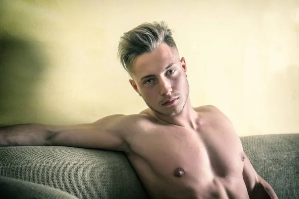 A shirtless man laying on a couch. Photo of a handsome blond man with muscular physique lounging on a couch looking at camera