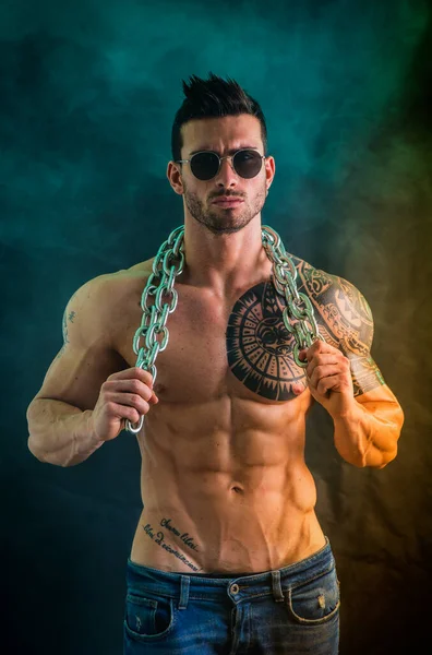 stock image Handsome shirtless muscular man with jeans, standing and wearing sunglasses, on dark smoky background in studio shot. Tattoo reads: We are free to start again, in Italian