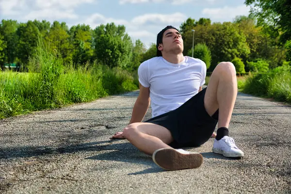 A man sitting on the side of a road. Photo of a young athlete taking a break on the side of a road during an outdoor workout
