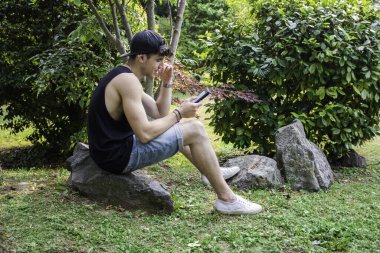 Çimenlerde bir kayanın üzerinde oturan bir adam, bir kitap okuyucudan kitap okuyor. Bir e-kitap okuyucusuyla çimlerin üzerinde oturan yakışıklı bir gencin fotoğrafı.