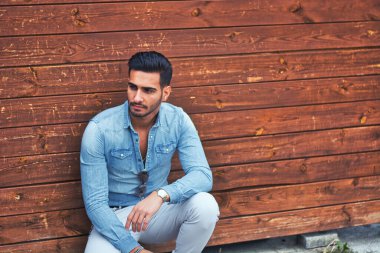 Ahşap bir duvarın önünde yerde oturan bir adam.