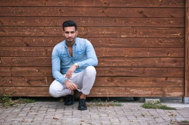 Ahşap bir duvarın önünde yerde oturan bir adam.