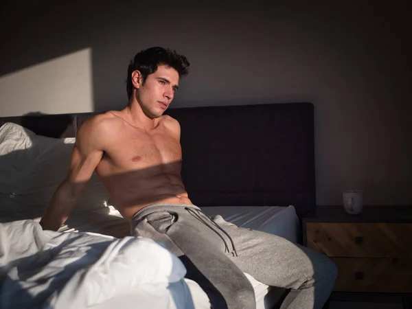 A shirtless man sitting on a bed in a bedroom. Photo of a shirtless man relaxing on a bed in the morning