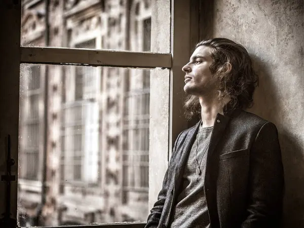 A man is looking out of a window. A young man with long hair looking out of a window