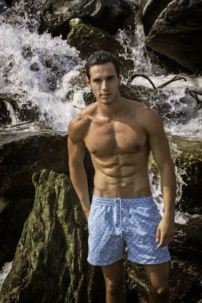A shirtless man standing in front of a waterfall.Photo of a young and attractive shirtless man posing in front of a majestic waterfall