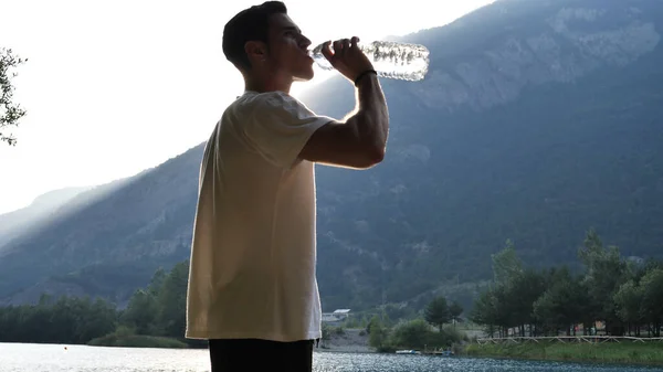 A man drinking water from a plastic bottle. Photo of a man quenching his thirst with a refreshing drink from a plastic water bottle, by a lake