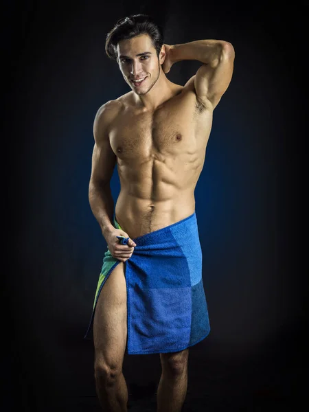 A young attractive man with a towel wrapped around his muscular body. Photo of a man wrapped in a towel in studio, on dark background, looking at camera