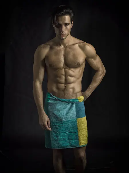 A young attractive man with a towel wrapped around his muscular body. Photo of a man wrapped in a towel in studio, on dark background, looking at camera
