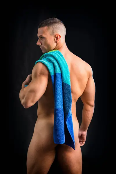 A shirtless man with a towel on his shoulder, covering his buttock. Portrait of naked handsome young man with languishing look covering crotch with a towel, against dark background