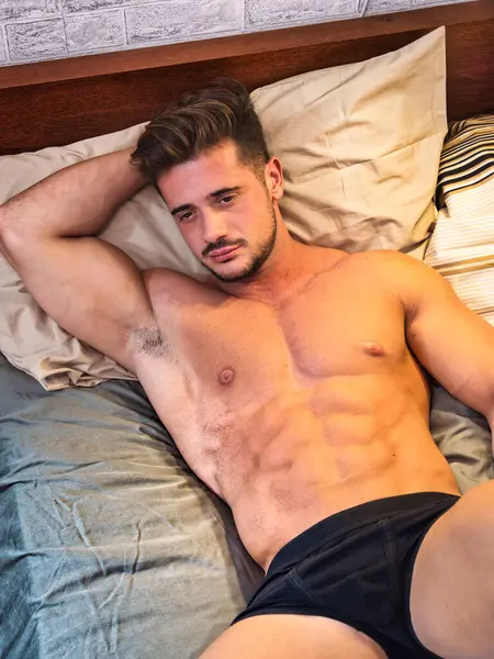 A muscular man laying on a bed with no shirt on. Photo of a shirtless man lying on a bed