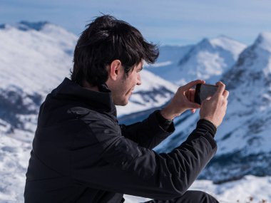 Karla kaplı bir dağın tepesinde oturan bir adam akıllı telefonuyla manzaranın fotoğrafını çekiyor.