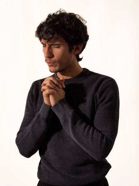 A young handsome man in a black sweater is praying, holding his hands joined together, isolated on white in studio