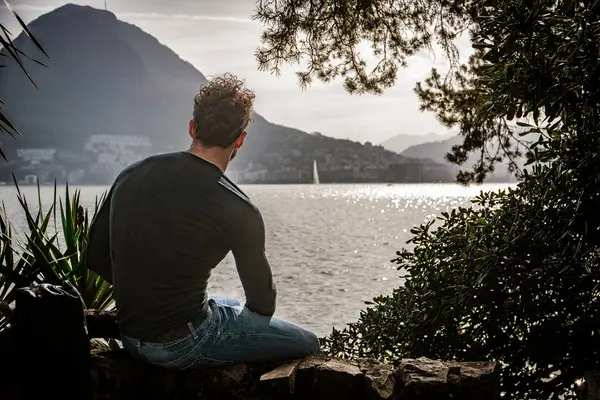 A man sitting on a rock looking out over a body of water in Lugano, Switzerland