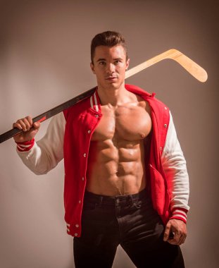 A muscular young man holding a hockey stick and wearing a red jacket open on muscle torso. A Strong Man Ready to playing hockey clipart