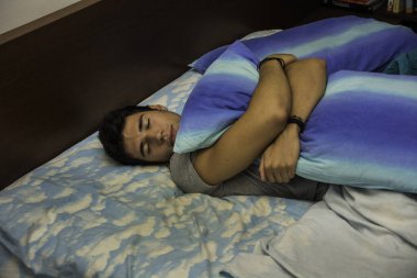 Yatakta uzanmış, mavi ve beyaz bir yastık tutan genç bir adam. Genç ve çekici bir adamın fotoğrafı. Yatakta uzanmış şık mavi beyaz bir yastığa sarılıyor.