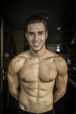 Genç, yakışıklı, kaslı, gömleksiz bir adam. Evin içinde fotoğraf için poz veriyor. Kameraya bakıp gülümsüyor.