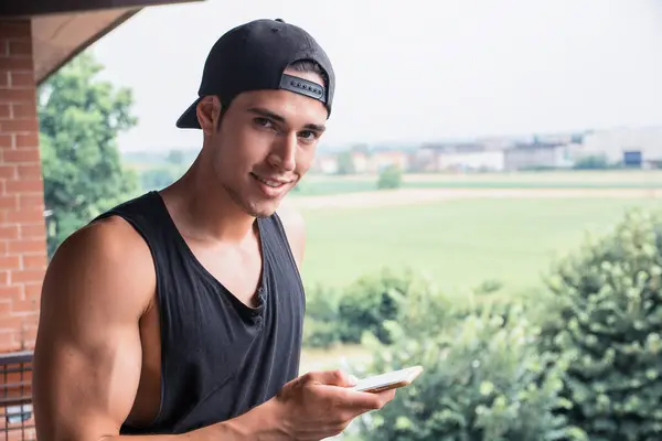 A man in a black tank top holding a cell phone, outdoor, looking at camera smiling