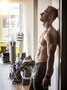 Yakışıklı, kaslı, üstsüz genç adam spor salonunda duruyor, antrenmandan sonra dinleniyor, önüne bakıyor, açık kapıya yaslanıyor.