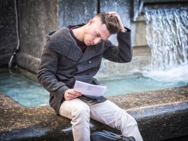 Genç adam, bir öğrenci, kağıt sayfalarının bir kış günü şehir ayarında okuma Çeşme başına oturmuş