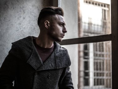 Eski binanın içinde, büyük pencerenin yanında duran, yün kazak giyen, dışarı bakan yakışıklı, ciddi bir adam.