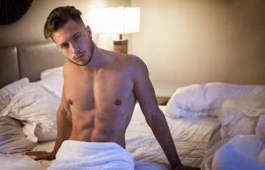 Yatak odasında, baştan çıkarıcı bir tavırla kameraya bakarak yatağında yalnız yatan gömleksiz seksi erkek modeli
