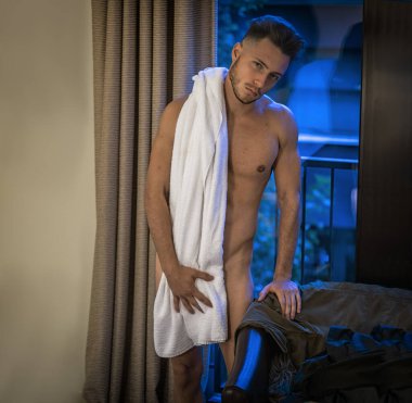Tamamen çıplak genç bir adam duş aldıktan sonra yatak odasında kameraya bakıyor, büyük bir havluyla üstünü örtüyor.