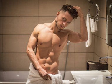 Gömleksiz, kaslı, yakışıklı genç bir adam sabah banyoda kameraya bakıyor, belinde sadece beyaz bir havlu var.