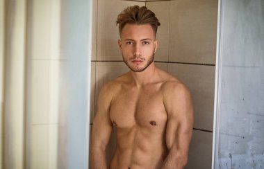 Çıplak Atletik Genç Adam, Cam Kapıya yaslanırken Dinlenme Banyosu 'nda duş alıyor, Kasıkları Elle Kaplıyor, Kameraya Bakıyor