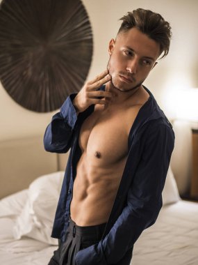 Seksi erkek model yatak odasında tek başına duruyor, baştan çıkarıcı bir tavırla kameraya bakıyor, kaslı göğüs ve gövdesinde gömleği açık.