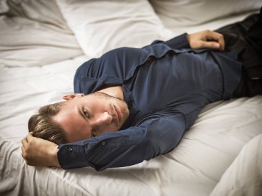 Seksi erkek model yatak odasında tek başına uzanıyor, baştan çıkarıcı bir tavırla kameraya bakıyor, mavi zarif bir gömlek giyiyor.