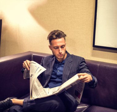 Evdeki yakışıklı genç adam gazete okuyor, deri koltukta oturuyor.