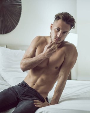 Yatak odasında, baştan çıkarıcı bir tavırla kameraya bakarak yatağında yalnız yatan gömleksiz seksi erkek modeli