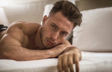 Yatak odasında, baştan çıkarıcı bir tavırla kameraya bakarak yatağında yalnız yatan gömleksiz seksi erkek modeli